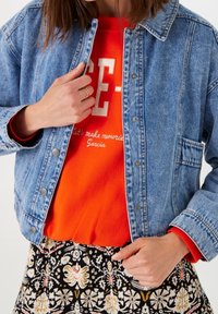 Modrá džínová bunda s kovovými patentními knoflíky, nošená přes oranžovou mikinu s bílým nápisem, doplněná květovanou sukní.