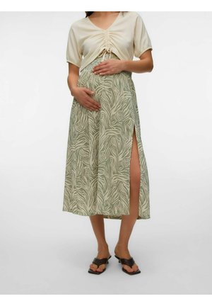 Femme enceinte portant un haut beige froncé et une jupe verte à imprimé zèbre avec une fente latérale, chaussures ouvertes à talons noires, mains sur le ventre.