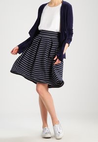 Gonna a righe blu e bianche con pieghe, abbinata a una blusa bianca e un cardigan blu; indossata con sneakers argentate. Tessuto dalla texture liscia.