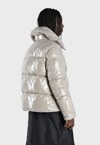 Doudoune en beige métallique, avec capuche, design rembourré avec sections matelassées et texture lisse et brillante.