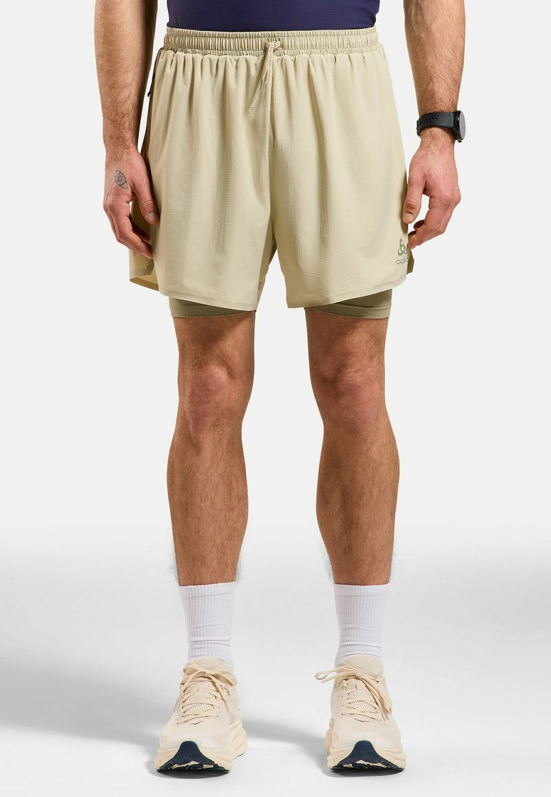 Helle beige Laufshorts mit einem elastischen Bund, die über eine Gitterstruktur und Seitentaschen verfügen, getragen über grünen Kompressionsshorts.