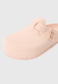 Roza gumena klompa s glatkom teksturom, otvorenim petom i jednim prilagodljivim remenom preko vrha. Ugrožena potplat za najbolji grip.