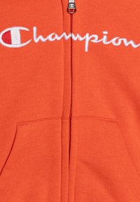 Hoodie naranja con cremallera hecho de tela cepillada. Presenta el logotipo "Champion" bordado en blanco y dos bolsillos frontales. Cremallera con tirador de metal.