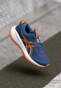 Μπλε αθλητικό παπούτσι με πορτοκαλί λεπτομέρειες, αναπνεύσιμο άνω μέρος από δίχτυ, καμπύλη λευκή σόλα και ανακλαστικές λεπτομέρειες στο πλάι. Ελαφριά σχεδίαση.