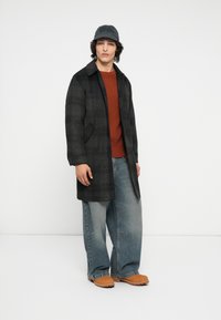 Sort plaid frakk, rød ribbet sweater og oversized blå jeans. Brune clogs og en sort kasket fuldender outfittet, som har en afslappet pasform.