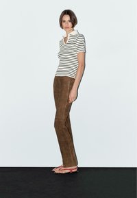 Randig kortärmad polo skjorta i vitt och svart, i kombination med bruna mockabyxor. Modellen bär platta röda sandaler. Enkel, figurnära design.