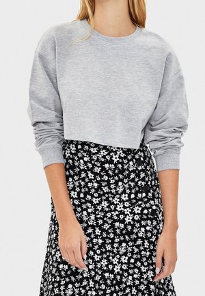 Femme portant un sweat-shirt court gris clair à manches longues et une jupe noire avec un motif floral blanc.