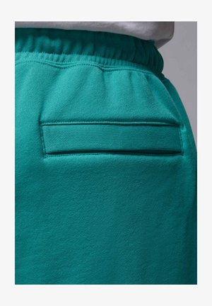Turquoise stoffen broek met elastische tailleband en een horizontale schuine zak aan de achterkant, gedragen met een wit bovenstuk.