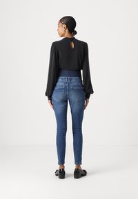 Black long-sleeve blouse with a keyhole back, paired with blue fitted jeans. The jeans feature a smooth finish and subtle back pocket stitching.