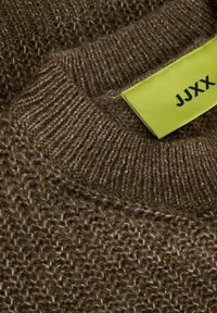 Hnědý pletený svetr zblízka, s texturovaným vzorem a ribbovaným límcem. Jasně zelená nálepka s nápisem "JJXX" je výrazně zobrazená.