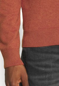 Vänster hand vid torso iförd en rostfärgad tröja och svarta jeans, som visar detaljerna på ärmslutet och tröjans nederkant.