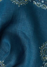 Blå linnetyg med intrikata guld- och vita blommönster längs kanten, med en mjuk textur och smidig drapering.