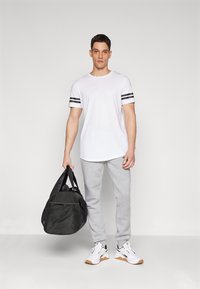 Hvit T-skjorte med kort erm og svarte striper, grå joggebukser og hvite sneakers med brune detaljer, holder en svart duffelbag.