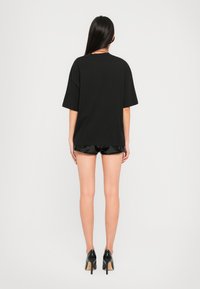 Black oversized t-shirt with short sleeves, back view. Paired with shiny black shorts and high-heeled shoes, against a plain background.