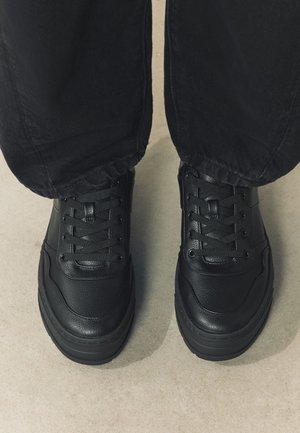 Chaussures noires à lacets portées avec un pantalon noir, vues de dessus sur un sol clair.