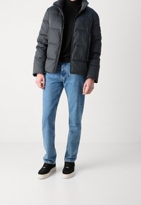Giubbotto a vento nero con texture trapuntata, chiusura con zip sul davanti e colletto alto, indossato sopra un dolcevita nero e jeans blu. Sneakers nere.