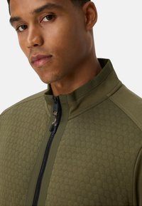 Olijfgroene ritsjack met een hexagonaal textuurpatroon, een glad bovenpaneel en een zwarte rits met een trekhaak.