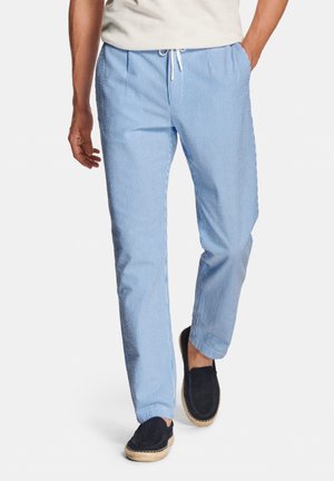 Man loopt in lichtblauwe en wit gestreepte broek met witte trekkoorden, wit shirt en zwarte espadrilles.