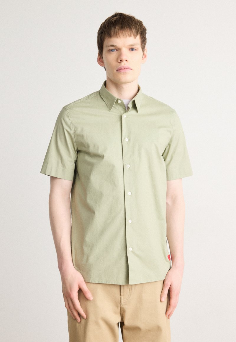 Camisa de botones de manga corta en algodón verde claro. Presenta un cuello, botones en el frente y una pequeña etiqueta roja en el lado.