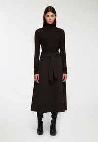 Maglione a collo alto nero e gonna lunga nera a vita alta con dettaglio di fiocco sul davanti; abbinata a stivali neri al ginocchio, texture liscia.