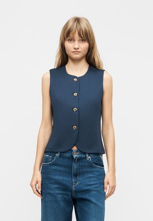 Jonge vrouw met steil blond haar, gekleed in een mouwloos marineblauw vest met knopen en een blauwe spijkerbroek, staand tegen een effen achtergrond.