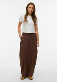 Witte cropped t-shirt, bruine broek met een ontspannen pasvorm en zakken, textuurstof, elastische tailleband, gedragen met beige instapschoenen.