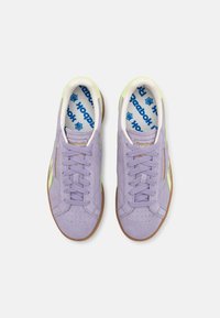 Baskets en daim violet clair avec accents verts, surface texturée, semelle en caoutchouc naturel, et logo Reebok bleu sur la semelle intérieure.