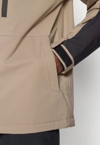 Το χέρι ενός ατόμου σε τσέπη μπεζ ζακέτας με μαύρο μανίκι που διαθέτει βέλκρο, σε λευκό φόντο.