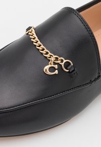 Mocassins en cuir noir avec un accent de chaîne dorée et un charme. Texture lisse, forme arrondie et détails de couture réguliers.