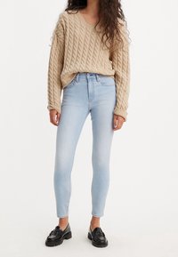 Suéter beige de punto trenzado con escote en V y corte holgado, combinado con jeans ajustados de color azul claro y mocasines negros.