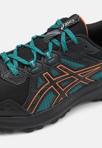 Chaussure de course noire avec une tige texturée, lacets teal avec des accents orange et panneaux en mesh. Semelle robuste conçue pour l'adhérence.