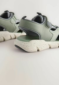 Zielone perforowane sneakersy z rzepami, z białą, cętkowaną podeszwą i mocną czarną podeszwą z przyczepnością. Widok z tyłu ukazuje wycięty design.
