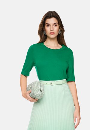 Frau mit langen braunen Haaren, die ein grünes kurzärmeliges Oberteil und einen hellgrünen plissierten Rock trägt, hält eine silberne strukturierte Clutch.