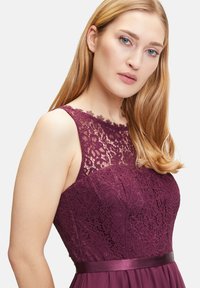 Bordeauxfarbiges Kleid mit einem Spitzen-Oberteil mit floralen Mustern, einem glatten Satin-Taillenbund und einem fließenden Rock. Der Ausschnitt ist gewellt.