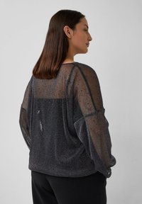 Durchscheinendes schwarzes Oberteil mit metallischen Fäden, langen Ärmeln und lockerem Schnitt. Auf der Rückseite befindet sich ein unifarbener Einsatz und eine leichte Raffung an der Schulter.