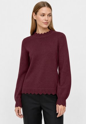 Femme portant un pull bordeaux à manches longues avec des bords festonnés et un pantalon noir, debout devant un fond clair uni.