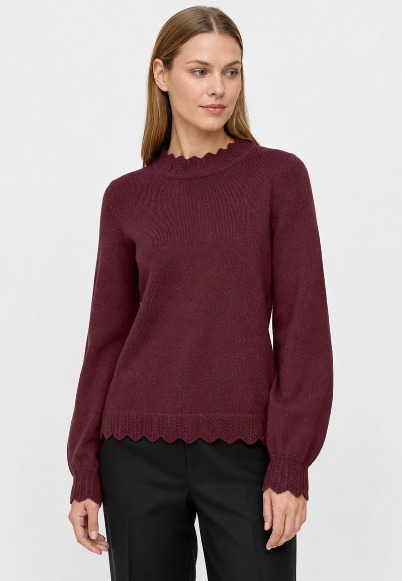 Femme portant un pull bordeaux à manches longues avec des bords festonnés et un pantalon noir, debout devant un fond clair uni.