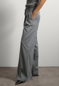 Grijze broek met wijde pijpen van een gladde stof, voorzien van plooien en zijzakken, gecombineerd met puntige zwarte schoenen op een neutrale achtergrond.