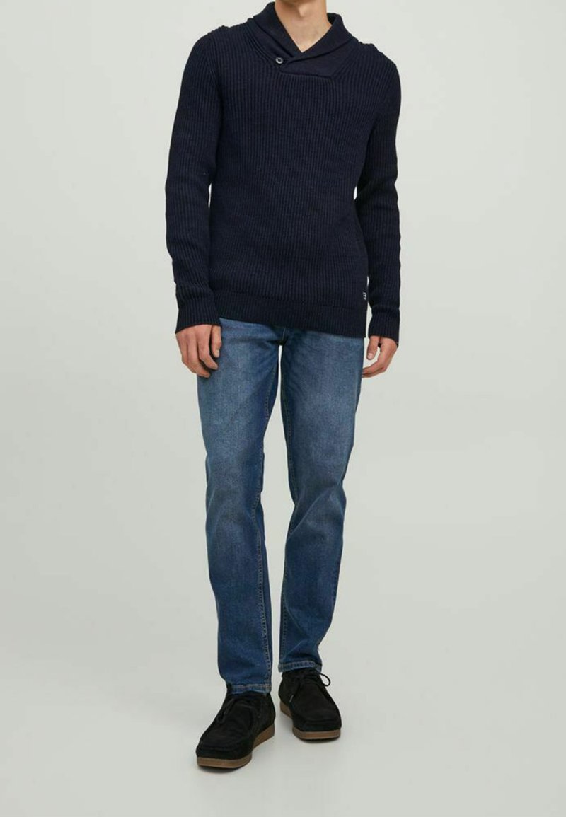 Homme portant un pull en maille bleu marine foncé avec col plié et bouton, jean bleu, et chaussures à lacets en daim noir, se tenant devant un fond uni.