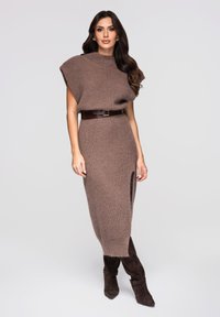 Robe en tricot marron à manches courtes, taille cintrée, fente latérale et ceinture marron. Portée avec des bottines en daim marron. Texture lisse.