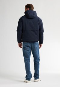 Geacă pufă navy cu glugă, țesătură texturată, buzunare cu fermoar, asociată cu blugi albastru deschis și adidași albe, văzută din spate.