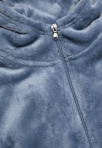Blå fleece zip-jacka med en mjuk, plyschig textur. Har en silverfärgad dragkedja och en huva. Tyget har en något glansig yta.