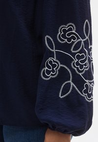 Blouse bleu marine à manches longues, ornée de motifs floraux blancs brodés et de poignets bouffants. Tissu lisse, légèrement texturé.