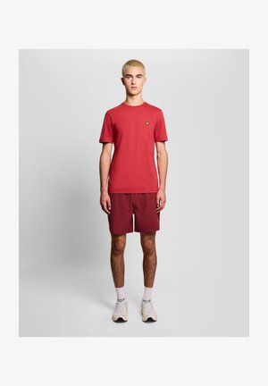 Ung mand med kort blondt hår iført en rød T-shirt, matchende røde shorts, hvide sokker og hvide sneakers, stående mod en ensfarvet baggrund.