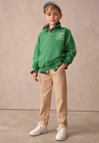 Sweat-shirt zippé vert superposé à une chemise à carreaux, pantalon beige et baskets blanches. Comprend un logo brodé et des manches retroussées.