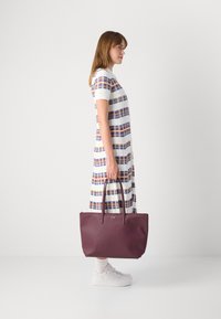 Γυναίκα με ριγωτό φόρεμα και λευκά αθλητικά παπούτσια κρατώντας μια μεγάλη μωβ τσάντα, στέκεται μπροστά από μια απλή λευκή επιφάνεια.