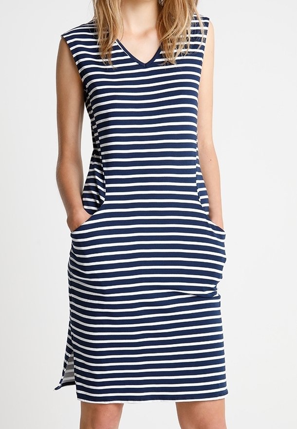 Kvinna klädd i ärmlös marinblå och vit horisontellt randig knälång klänning med V-ringning och fickor, stående med händerna i fickorna.