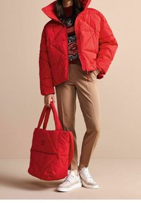 Giubbotto imbottito trapuntato rosso con colletto alto, abbinato a pantaloni beige su misura e sneakers bianche. Borsa tote rossa imbottita con dettagli di cucitura visibili.