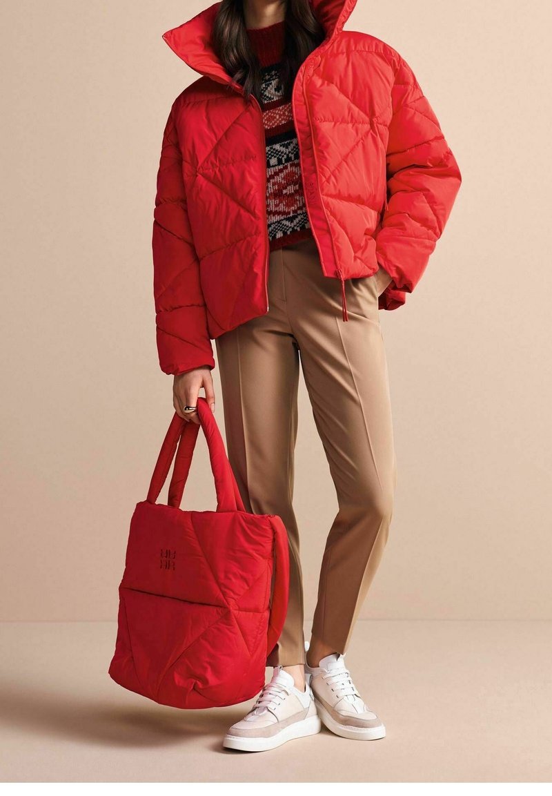 Giubbotto imbottito trapuntato rosso con colletto alto, abbinato a pantaloni beige su misura e sneakers bianche. Borsa tote rossa imbottita con dettagli di cucitura visibili.