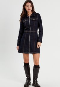 Vestido de mezclilla en azul oscuro con una silueta ajustada, mangas largas, cuello puntiagudo, cierre frontal con cremallera y costuras en un tono más claro.